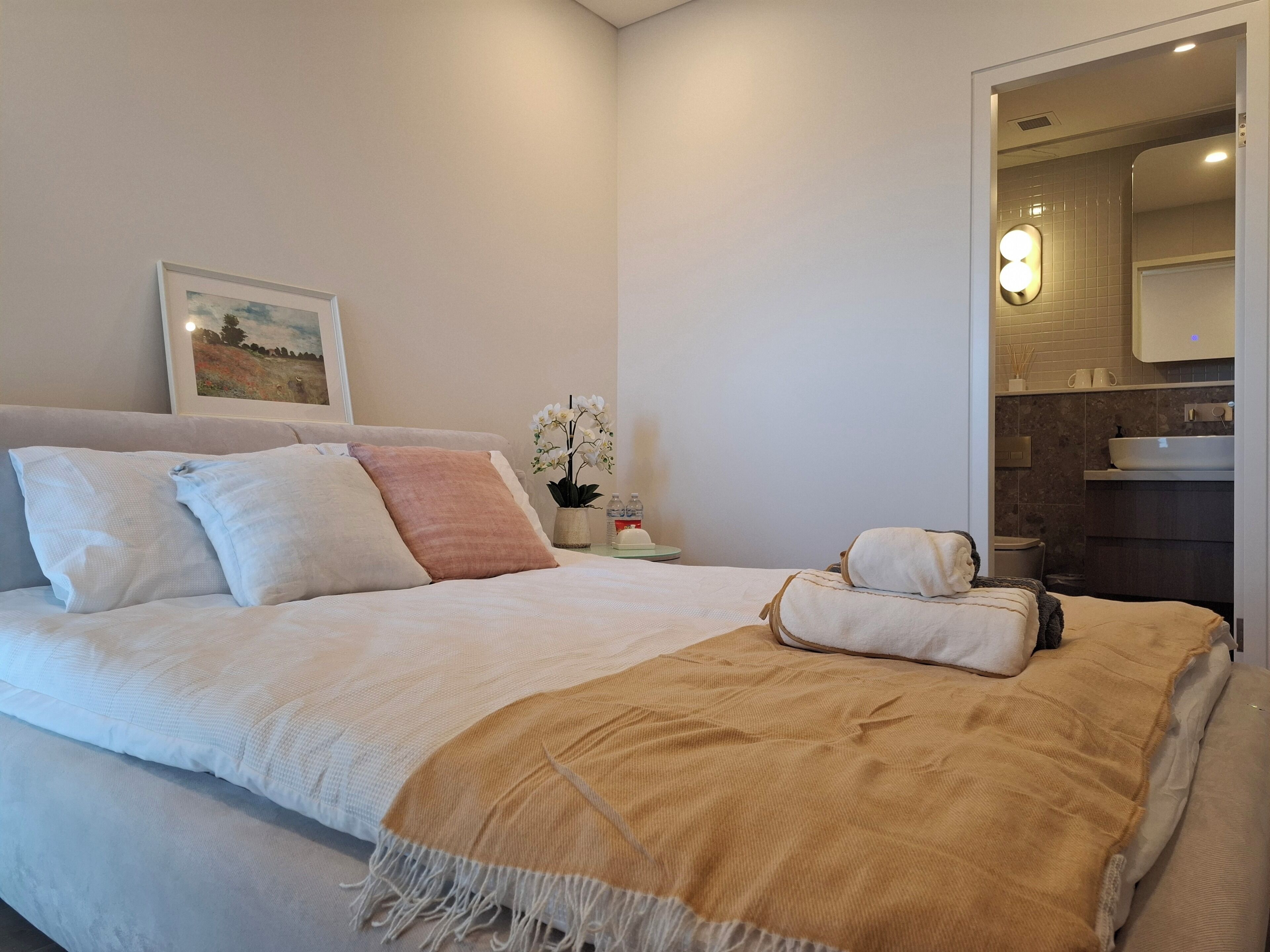A neatly made bed with pillows and a blanket, a bedside table with a plant and a bottle, and a bathroom visible through an open door.