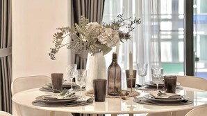 Een eettafel gedekt met borden, bestek en glazen, omringd door stoelen, met een vaas bloemen en een kaars als middelpunt.