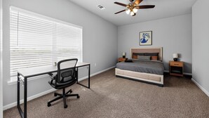 Quarto com cama, escrivaninha com cadeira, ventilador de teto, janela com persianas e obra de arte pendurada na parede.