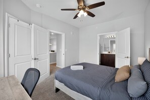 A bedroom with a bed, a desk, a chair, a ceiling fan, and a bathroom with a mirror and sink. - Comfortable 2BR Retreat (Princeton)