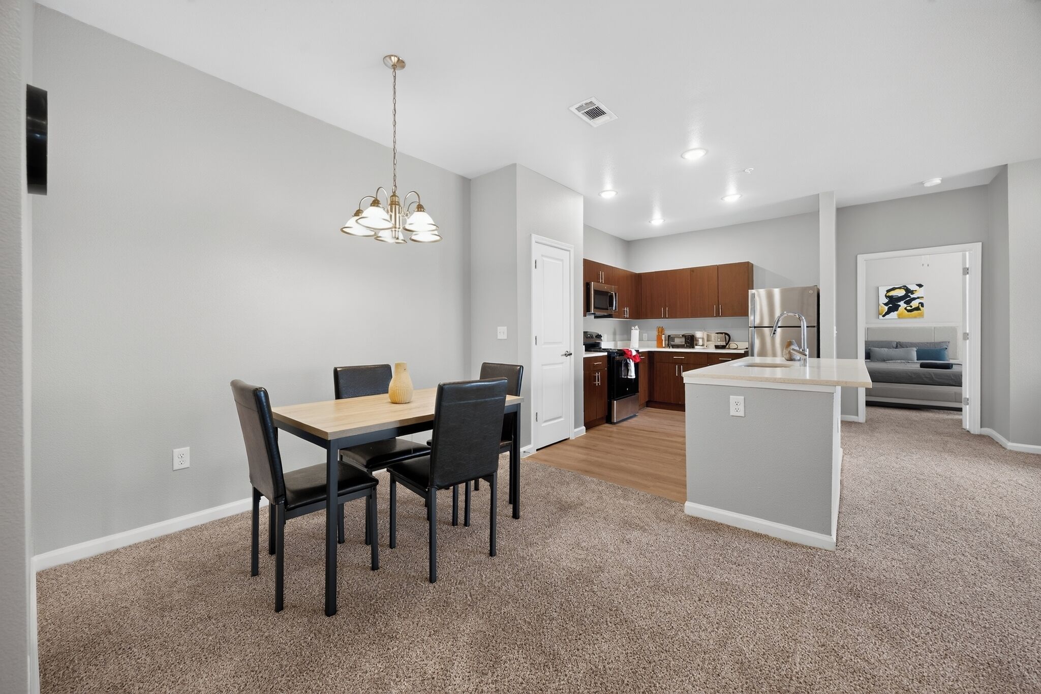 A modern kitchen with stainless steel appliances, a dining area with a wooden table and chairs, and a living area with a bed and artwork on the wall.