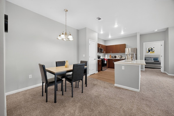 A modern kitchen with stainless steel appliances, a dining area with a wooden table and chairs, and a living area with a bed and artwork on the wall.