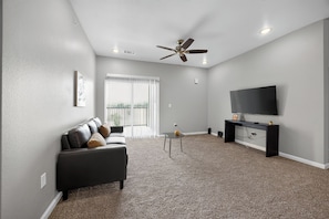 A living room with a sofa, a TV, a ceiling fan, and a balcony.