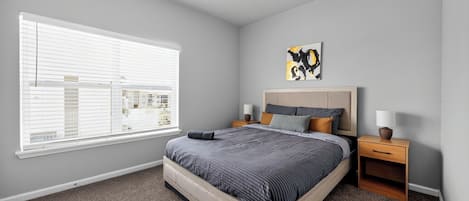 A bedroom with a large bed, a nightstand, a lamp, a window with blinds, and a ceiling vent.