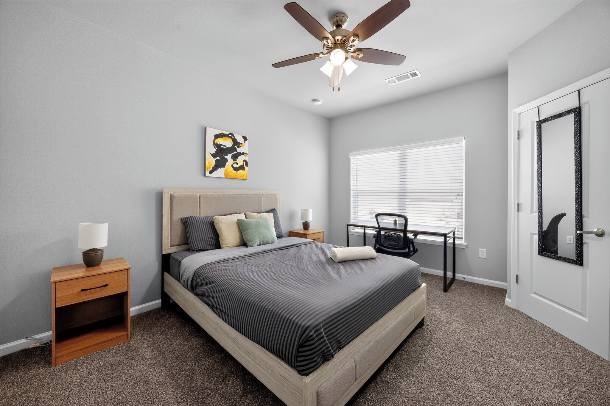 A bedroom with a bed, bedside tables, a ceiling fan, a window with blinds, and a door with a mirror.