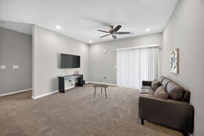 A modern living room with a flat-screen TV, a ceiling fan, a sofa, and a window with blinds. - Whitewing Flats Spacious 2BR (Princeton)
