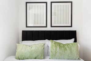 Een bed met een zwart hoofdeinde en twee kussens, een witte muur, en twee ingekaderde afbeeldingen erboven.