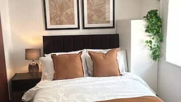 A hotel room with a bed, a nightstand, a lamp, framed artwork on the wall, and a window with a plant.
