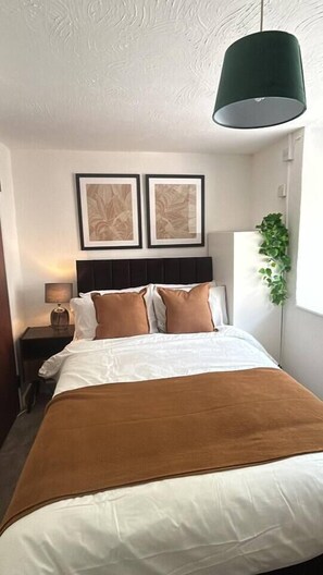 A hotel room with a bed, a nightstand, a lamp, framed artwork on the wall, and a window with a plant.