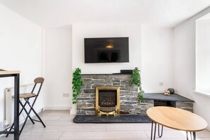 A modern living room with a stone fireplace, a flat-screen TV mounted above it, a dining table, and a chair. - #161 Stylish Bodmin Cornwall Stay - Sleeps 6 (Cornwall)