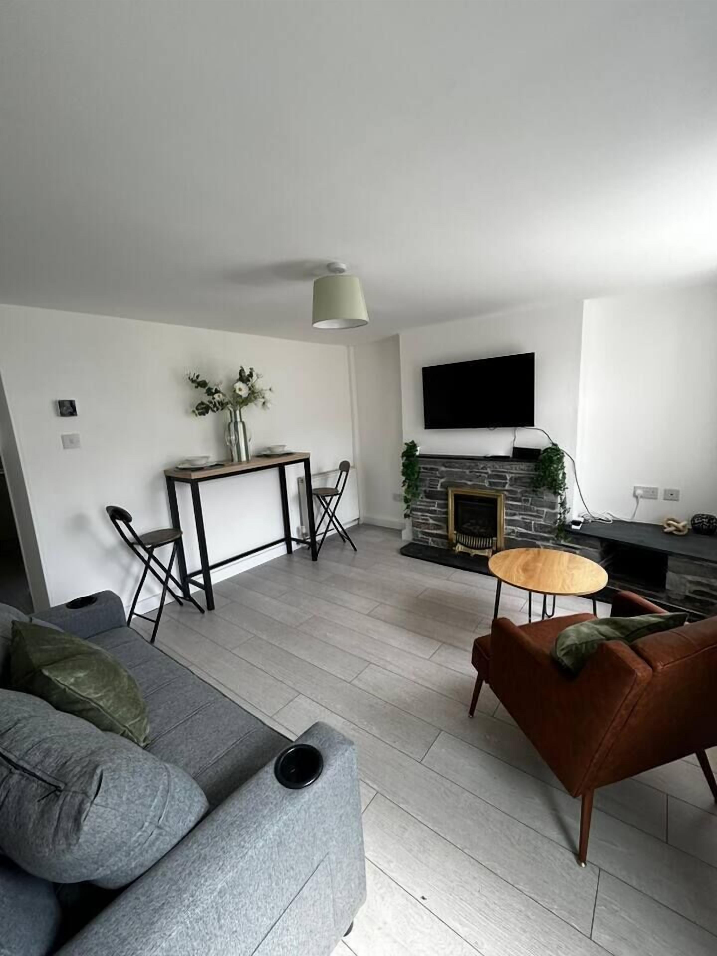 Een moderne woonkamer met een grijze bank, een bruine fauteuil, een klein rond tafeltje en een open haard met een stenen omlijsting.