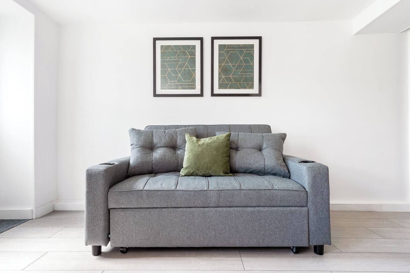 A grey sofa with a single green pillow in a room with white walls and two framed abstract art pieces above it.