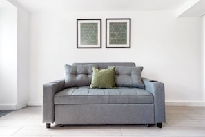 A grey sofa with a single green pillow in a room with white walls and two framed abstract art pieces above it.