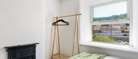 Ein Schlafzimmer mit einem Bett, einem Holzkleiderständer, einer schwarzen Kommode und einem Fenster mit Blick auf Gebäude.