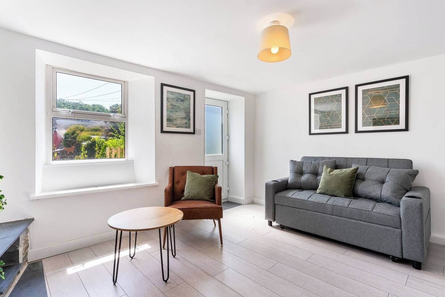 Ein modernes Wohnzimmer mit einem grauen Sofa, einem braunen Sessel und einem runden Tisch. Zwei Fenster bieten Blick ins Grüne, und eine Tür ist ebenfalls zu sehen.