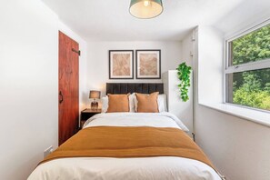 A bedroom with a bed, a nightstand, a wall-mounted lamp, two framed pictures, a window with a view of greenery, and a door with a handle. - #161 Stylish Bodmin Cornwall Stay - Sleeps 6 (Cornwall)
