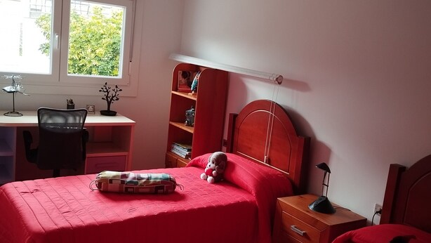 Ein Schlafzimmer mit zwei Betten, einem Schreibtisch, einem Stuhl, einem Bücherregal und einem Fenster mit Blick auf Grünflächen.