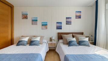 A hotel room with two beds, a wooden wardrobe, a nightstand with a lamp, and wall art depicting beach scenes.