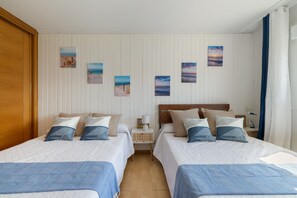 A hotel room with two beds, a wooden wardrobe, a nightstand with a lamp, and wall art depicting beach scenes.
