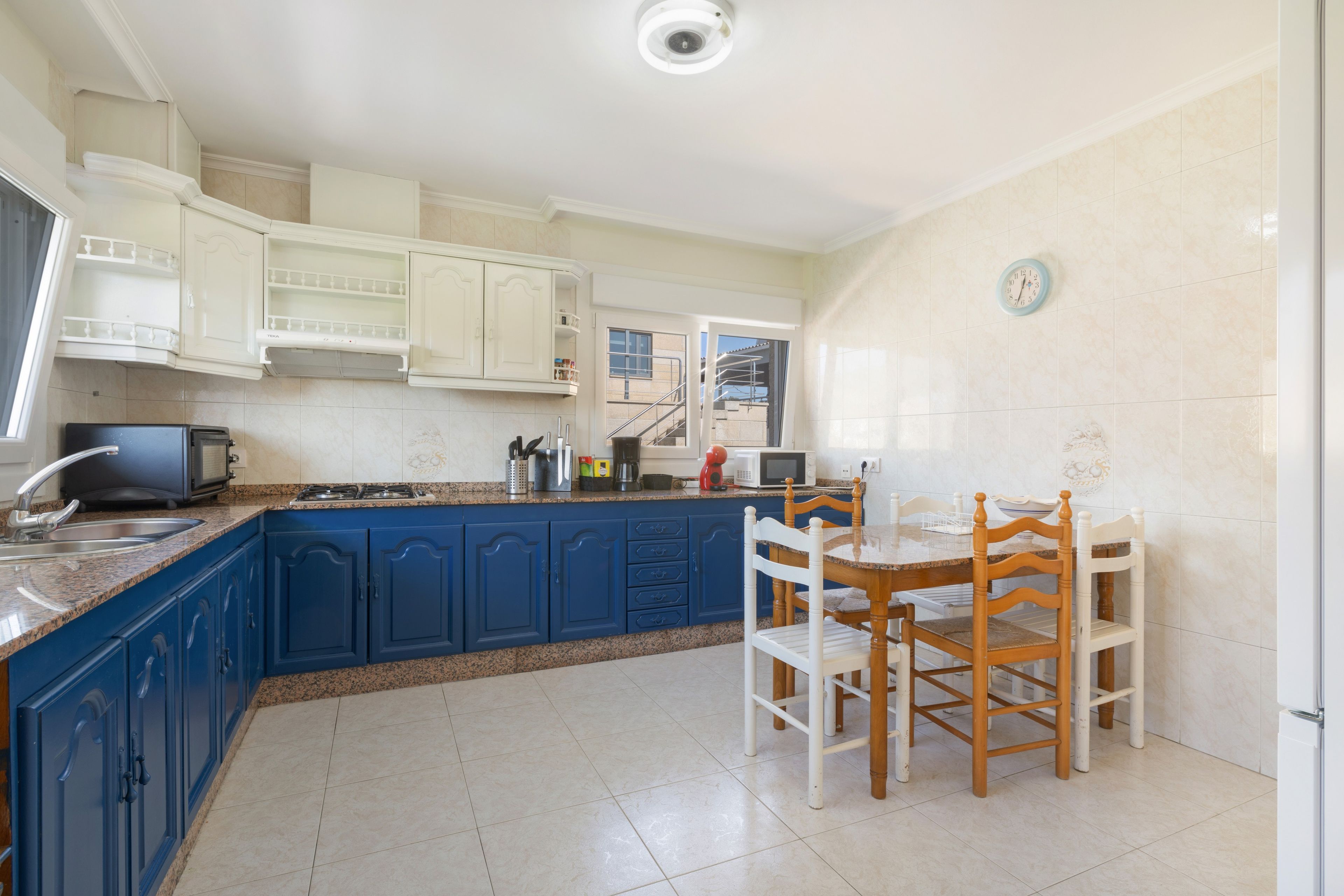 Une cuisine avec des meubles de rangement bleus, une table à manger avec des chaises, un four à micro-ondes et une horloge au mur.