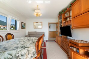 Un dormitorio con cama, televisor, aparador de madera y ventana con vista a casas.