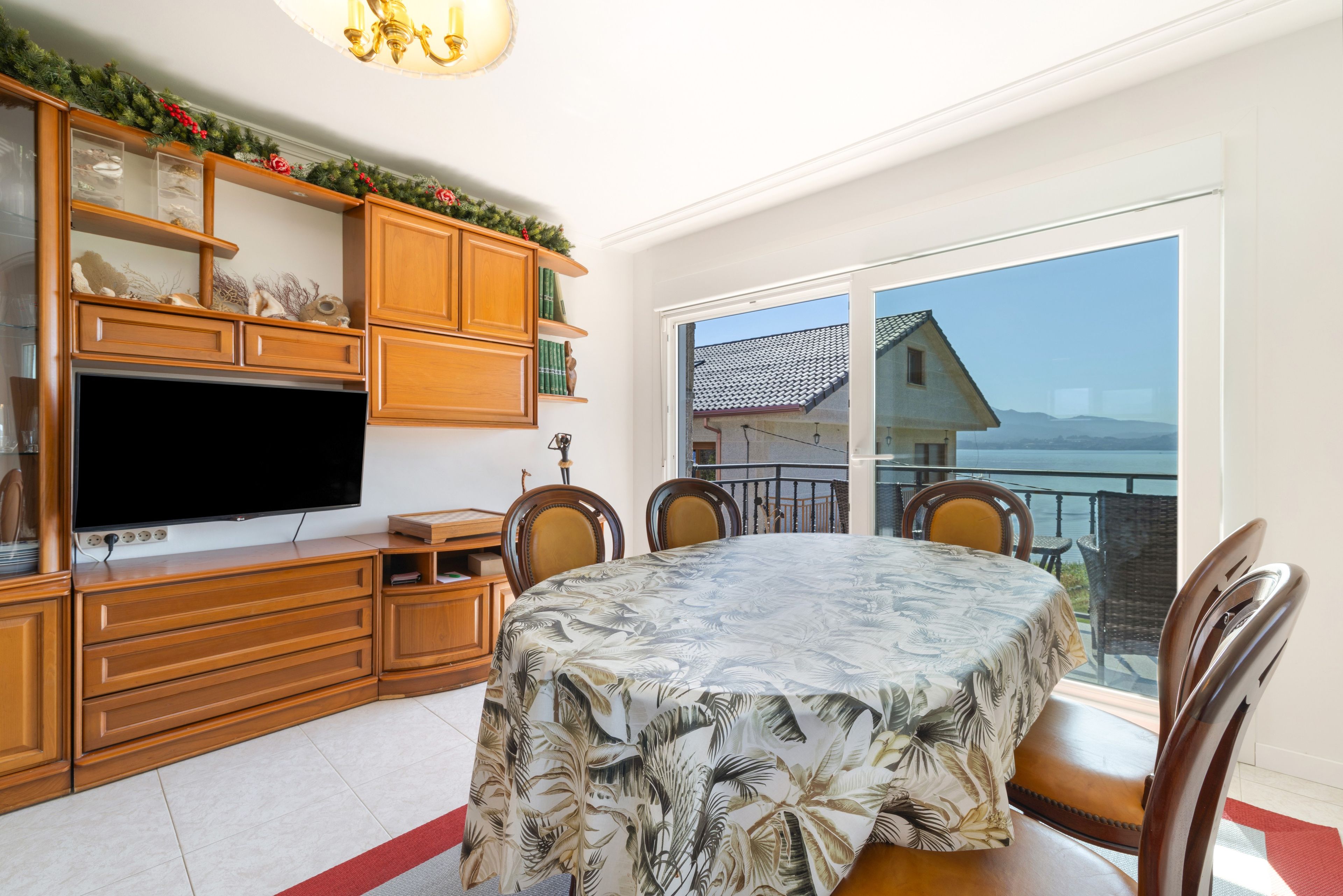 A dining area with a table covered in a tropical leaf tablecloth, wooden cabinets, a flat-screen TV, and a view of a house through a large window.