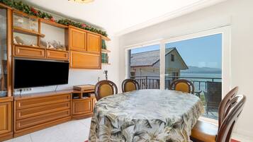 A dining area with a table covered in a tropical leaf tablecloth, wooden cabinets, a flat-screen TV, and a view of a house through a large window.