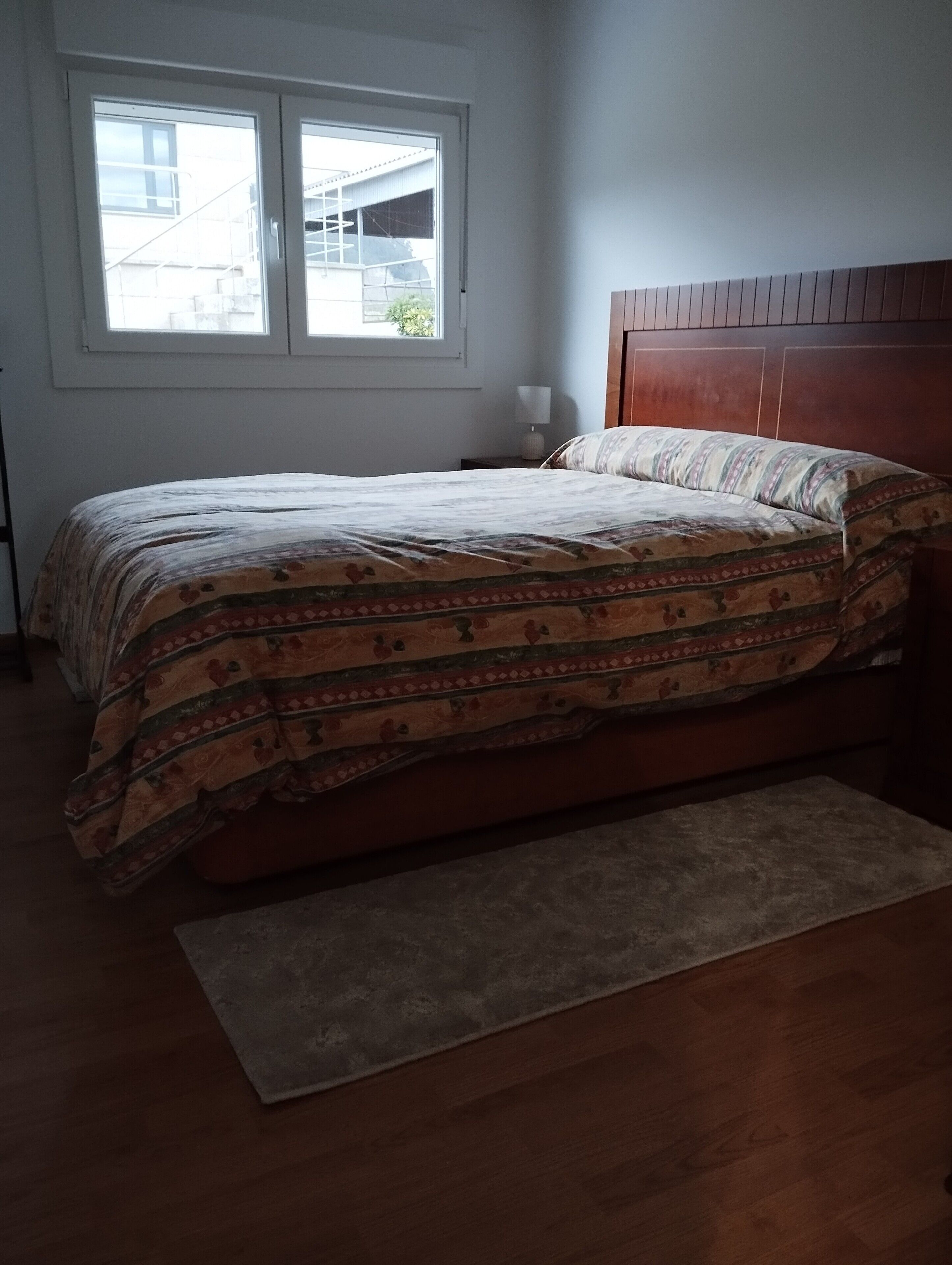 Ein Schlafzimmer mit einem Bett, einem Nachttisch, einer Lampe, einem Fenster und einem Holz-Kopfteil.
