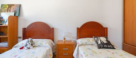 A bedroom with two single beds, a nightstand with a lamp, a shelf with books, and a wall-mounted painting.
