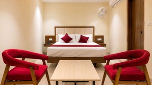 A hotel room with a bed, two red chairs, a small table, an air conditioning unit, and a wooden door. - Royal Suites in Marathalli Bangalore (Bengaluru)