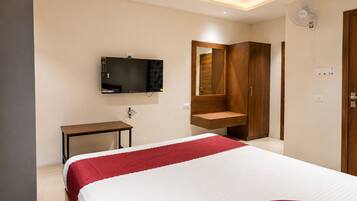 A hotel room with a bed, a TV mounted on the wall, a wooden wardrobe, and a small table.