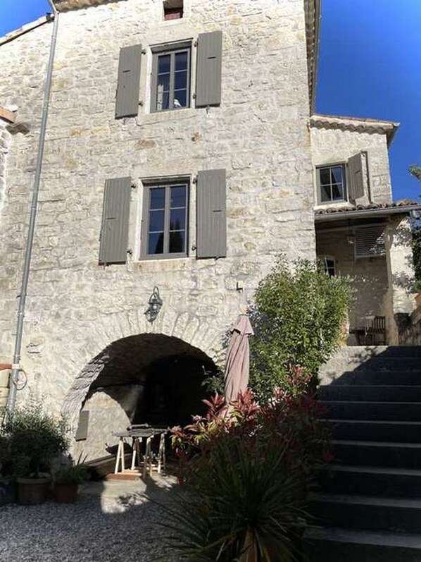 A stone building with a staircase leading to an upper level, a small archway entrance, and a garden area with plants and a table.