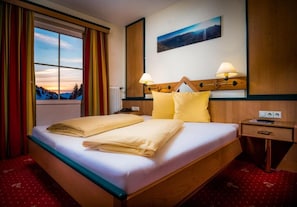 A hotel room with a bed, two bedside lamps, a nightstand, a window with curtains, and a painting on the wall. - Pension Weinberger (Obertauern)