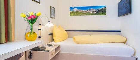 A single bed with a white mattress and yellow pillows, a bedside table with a lamp, a vase with flowers, and a picture of a mountain landscape on the wall.