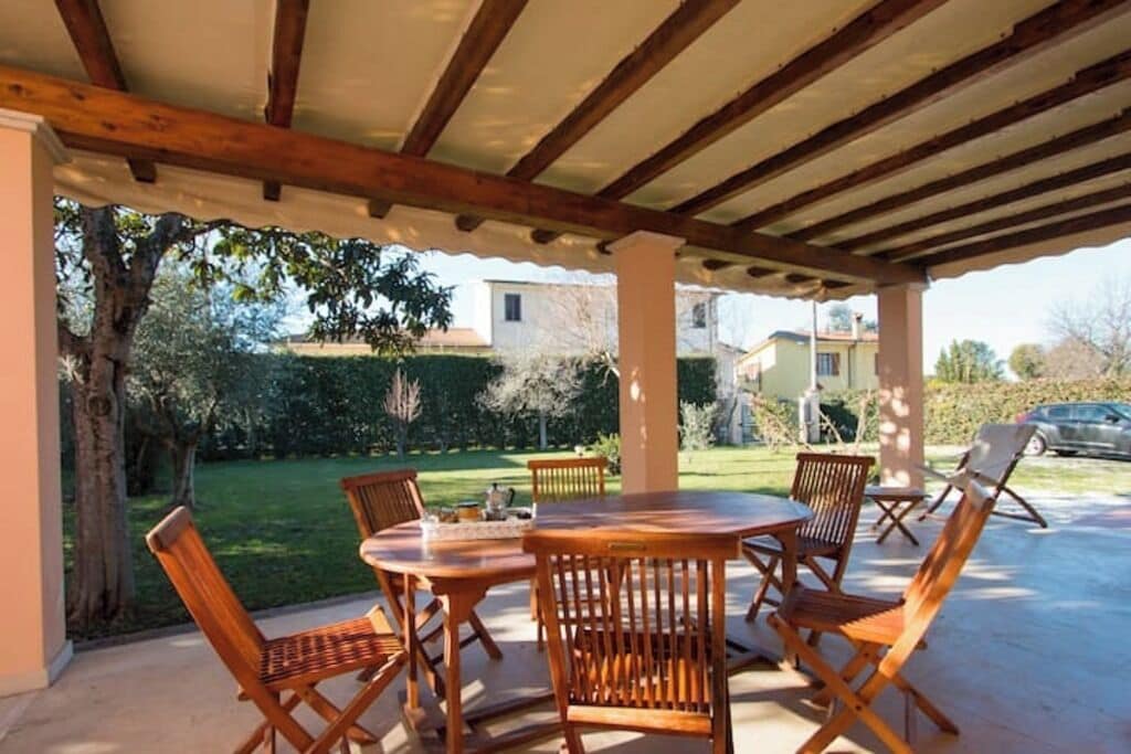 An outdoor patio with a wooden table and chairs, surrounded by a covered area with a view of a garden and houses in the background.