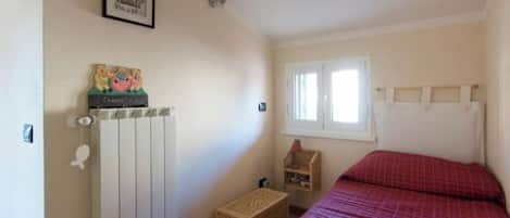 A bedroom with a bed, a radiator, a window, and a small wooden shelf.