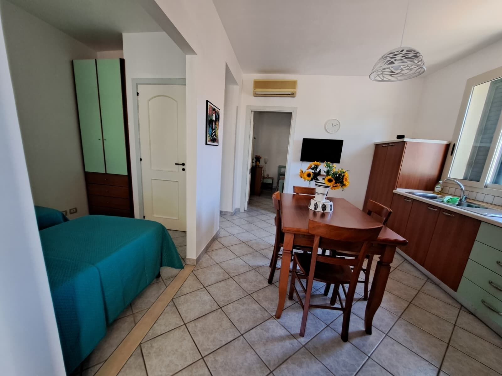 A compact room with a bed, a dining table with chairs, a kitchen area with a sink, and a television mounted on the wall.