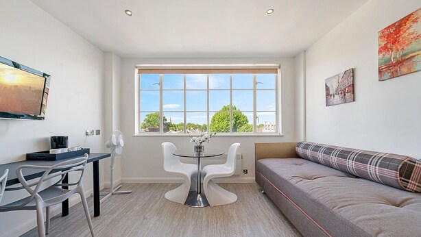 Een moderne woonkamer met een bank, een eettafel met stoelen, een groot raam met uitzicht op bomen en een schilderij aan de muur.