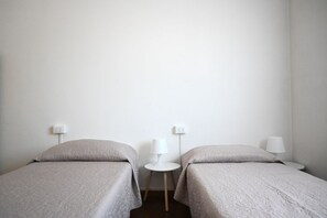 Quarto com duas camas de solteiro, cada uma com mesa de cabeceira e luminária, e uma tomada na parede.