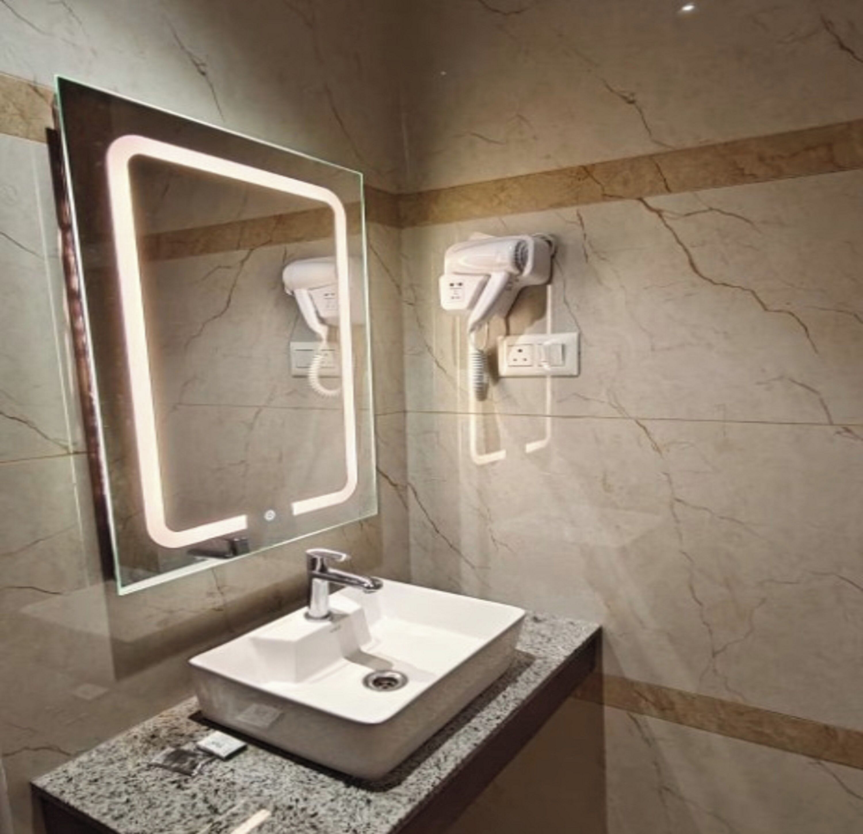 A bathroom with a marble wall, a sink with a modern faucet, a mirror with integrated lighting, and a hairdryer mounted on the wall.