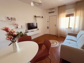 Ein modernes Wohnzimmer mit einem weißen Sofa, einem Esstisch mit Stühlen, einem Flachbildfernseher auf einer weißen Konsole und einem großen Fenster mit Vorhängen.
