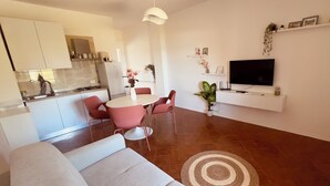 En moderne stue med sofa, spisebord med stole, fjernsyn og et køkken med skabe og køleskab.