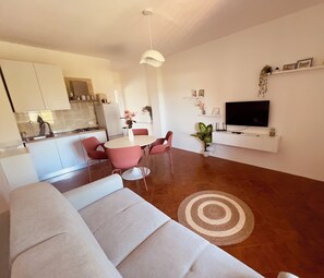 Ein modernes Wohnzimmer mit Sofa, Esstisch mit Stühlen, Fernseher und Küchenzeile mit Schränken und Kühlschrank.
