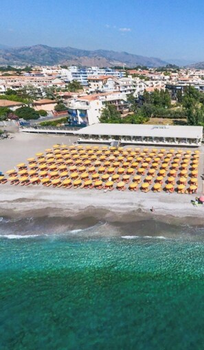 Ein Luftbild eines Strandes mit zahlreichen gelben Sonnenschirmen, die in Reihen angeordnet sind, einem klaren, blauen Meer und Gebäuden im Hintergrund.