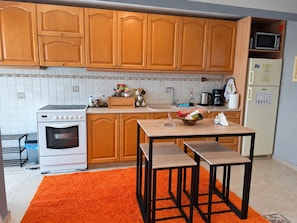 A kitchen with wooden cabinets, a white oven, a microwave, and a refrigerator. There is a table with chairs and a rug. - Aegean Sea & Mount Olympus View Guesthouse (Palaio Eleftherochori)