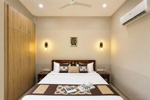 Une chambre d’hôtel comprenant un lit, des tables de chevet, une armoire, un ventilateur de plafond, des lampes fixées au mur et un appareil de climatisation.