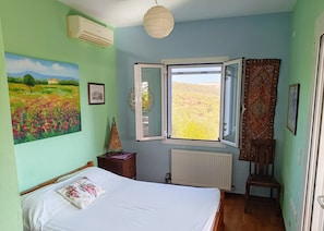 Un dormitorio con cama, radiador, ventana con vistas a un campo y un cuadro en la pared.