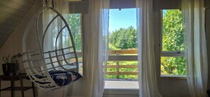 A room with a hanging chair, a window with a view of a wooden deck and greenery, and a door leading outside.