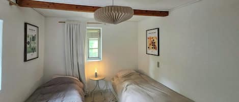 A bedroom with two single beds, a window with a curtain, a hanging light fixture, and two framed pictures on the walls.