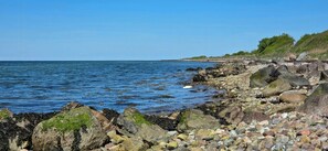 A rocky shoreline with clear blue water, a grassy hill in the background, and a clear sky. - Views across the fields to the sea on Langeland (Tranekær)